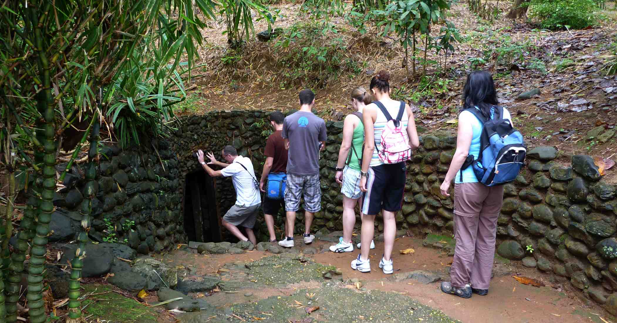 Nhóm khách du lịch cúi người bước vào cửa hầm địa đạo Vịnh Mốc, Quảng Trị, giữa rừng tre xanh